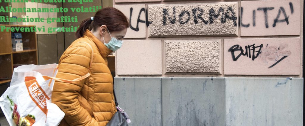 A woman wearing a protective mask walks next an inscription painted on the wall of a building reading in italian "normality", Genoa, Italy, 21 April 2020. Italian Prime Minister Giuseppe Conte, during his speech to the Chambers, said that the masks and the social security distancing will be maintained until a vaccine against Covid-19 wil not be discovered. ANSA/LUCA ZENNARO
