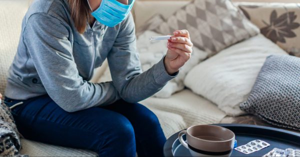 Ill infected woman checking thermometer having fever. Girl wearing protective mask while having cold, flu taking pills at home. Healthcare