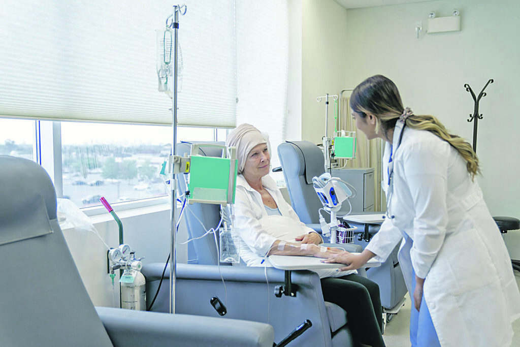 A senior woman is sitting on a chair in a chemotherapy treatment room. She is wearing a headscarf due to hair loss. Her female oncologist is checking in on her and providing her with moral encouragement.