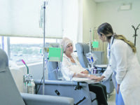 A senior woman is sitting on a chair in a chemotherapy treatment room. She is wearing a headscarf due to hair loss. Her female oncologist is checking in on her and providing her with moral encouragement.