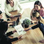 Businesswoman in group meeting discussion with other businesswomen colleagues in modern workplace office with laptop computer and documents on table. People corporate business working team concept.